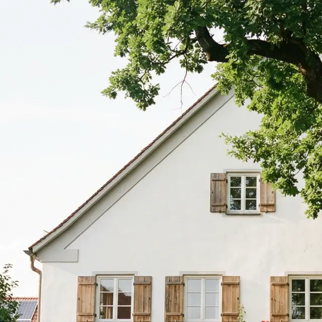 Ein großes, gepflegtes, traditionelles Haus mit hellen Wänden und Fensterläden. Davor steht ein großer, alter Baum in einem idyllischen Vorgarten.