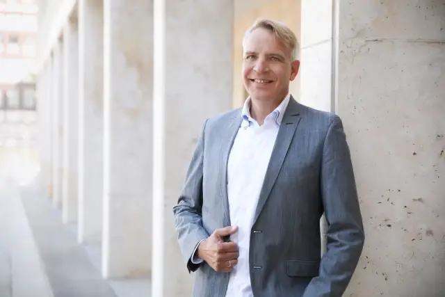 Porträt eines lächelnden, sympathischen Immobilienmaklers in einem grauen Sakko, der vor einem hellen, säulenartigen Hintergrund steht.