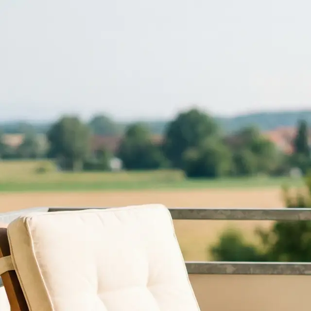 Ein bequemer Holzsessel mit einer hellen Decke steht auf einem Balkon. Im Hintergrund ist eine weite, grüne Landschaft mit einem Kirchturm zu sehen, was eine ruhige und entspannte Atmosphäre vermittelt.
